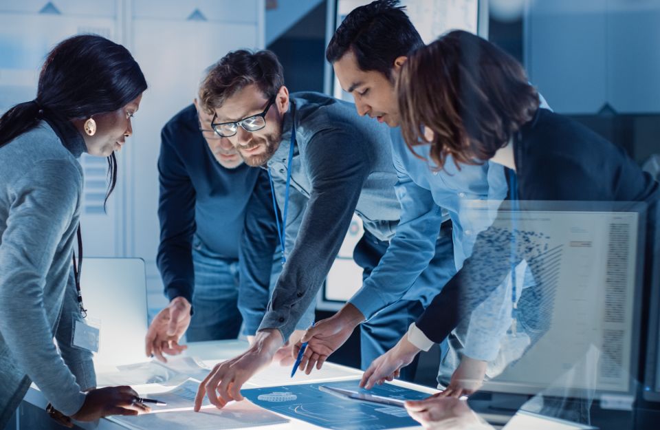 5 coworkers inspecting blueprints laid out across a desk